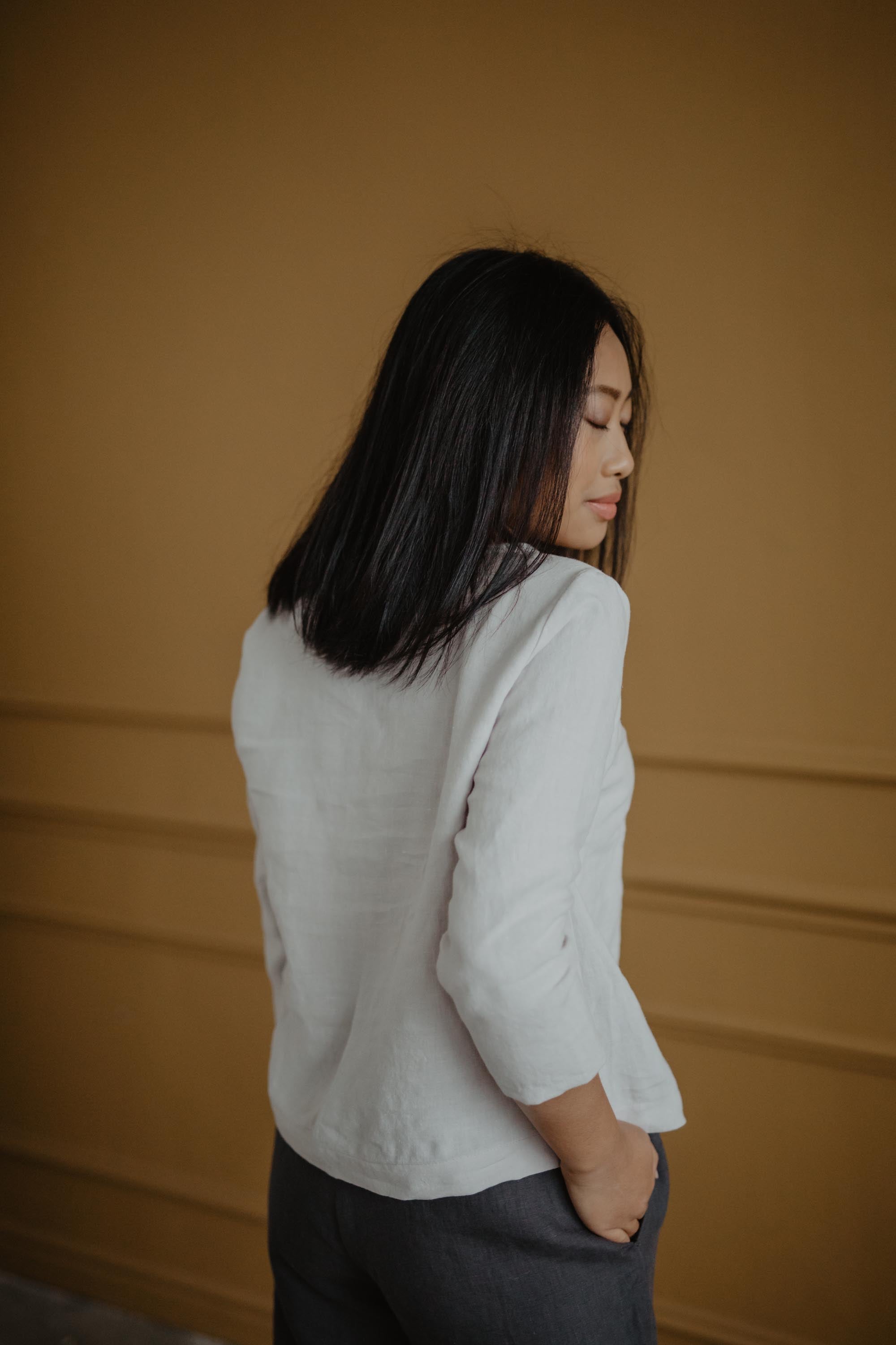Woman Looking OVer Shoulder Wearing A White Linen Top And Grey Linen Pants