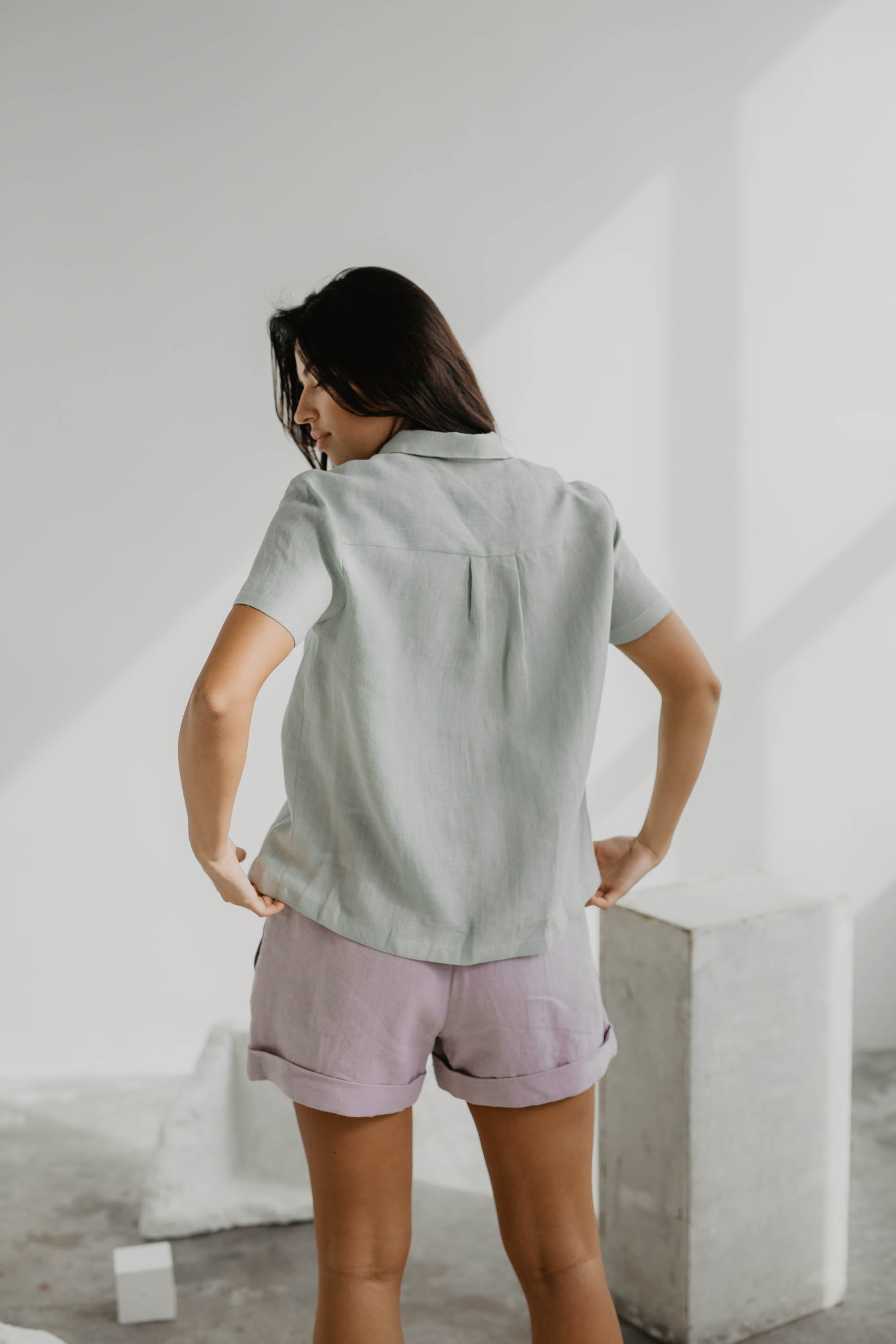 Woman Turned Away Wearing A Sage Green Linen Summer Shirt and Dusty Rose Linen Shorts