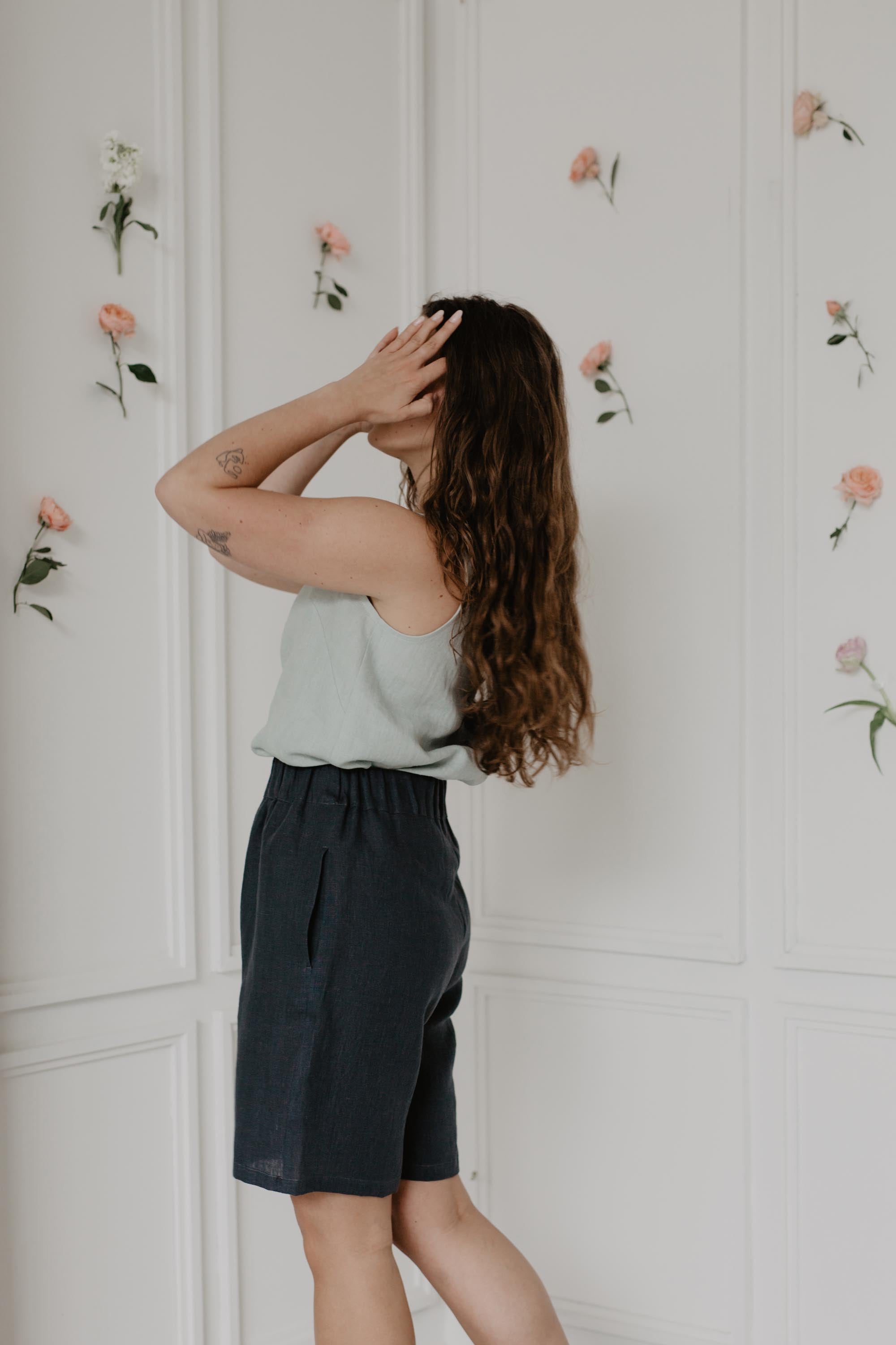 Woman Adjusting Hair Wearing Dark Linen Shorts and Sage Green Top In a Flowery Light Room