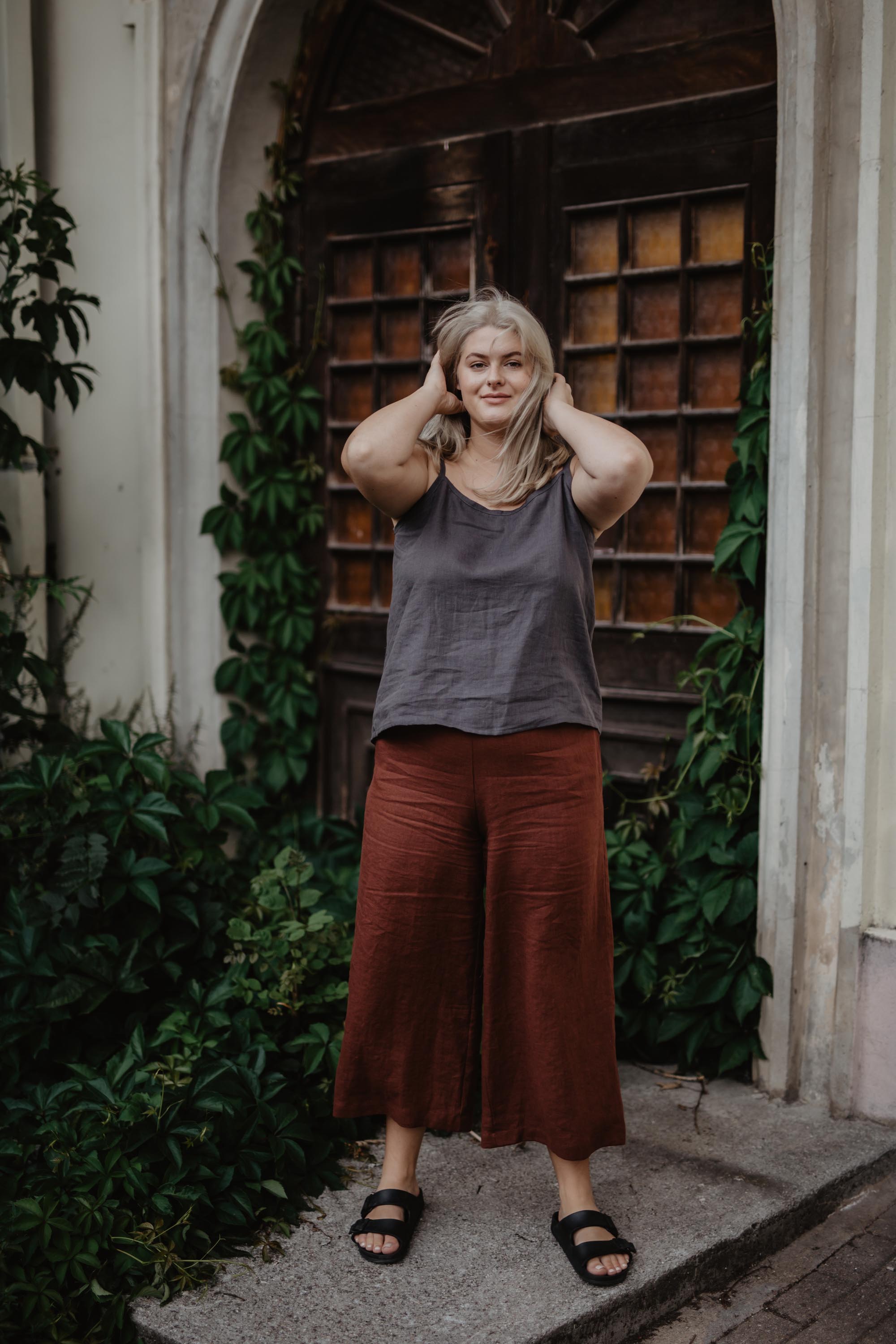 Woman In Botanical Setting Wearing Terracotta Linen Pants and Dark Linen Top By Amourlinen