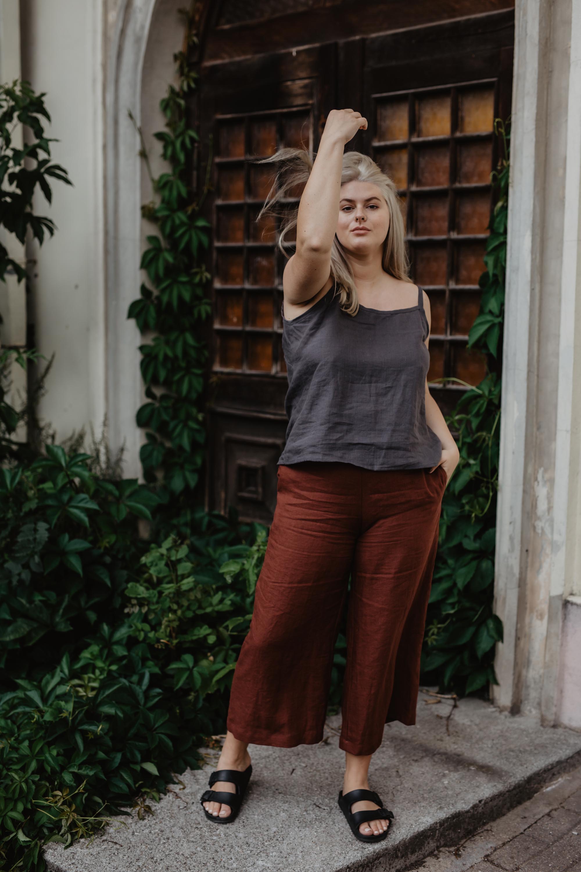 Woman In Botanical Setting Posing Wearing Terracotta Linen Pants and Dark Linen Top By Amourlinen