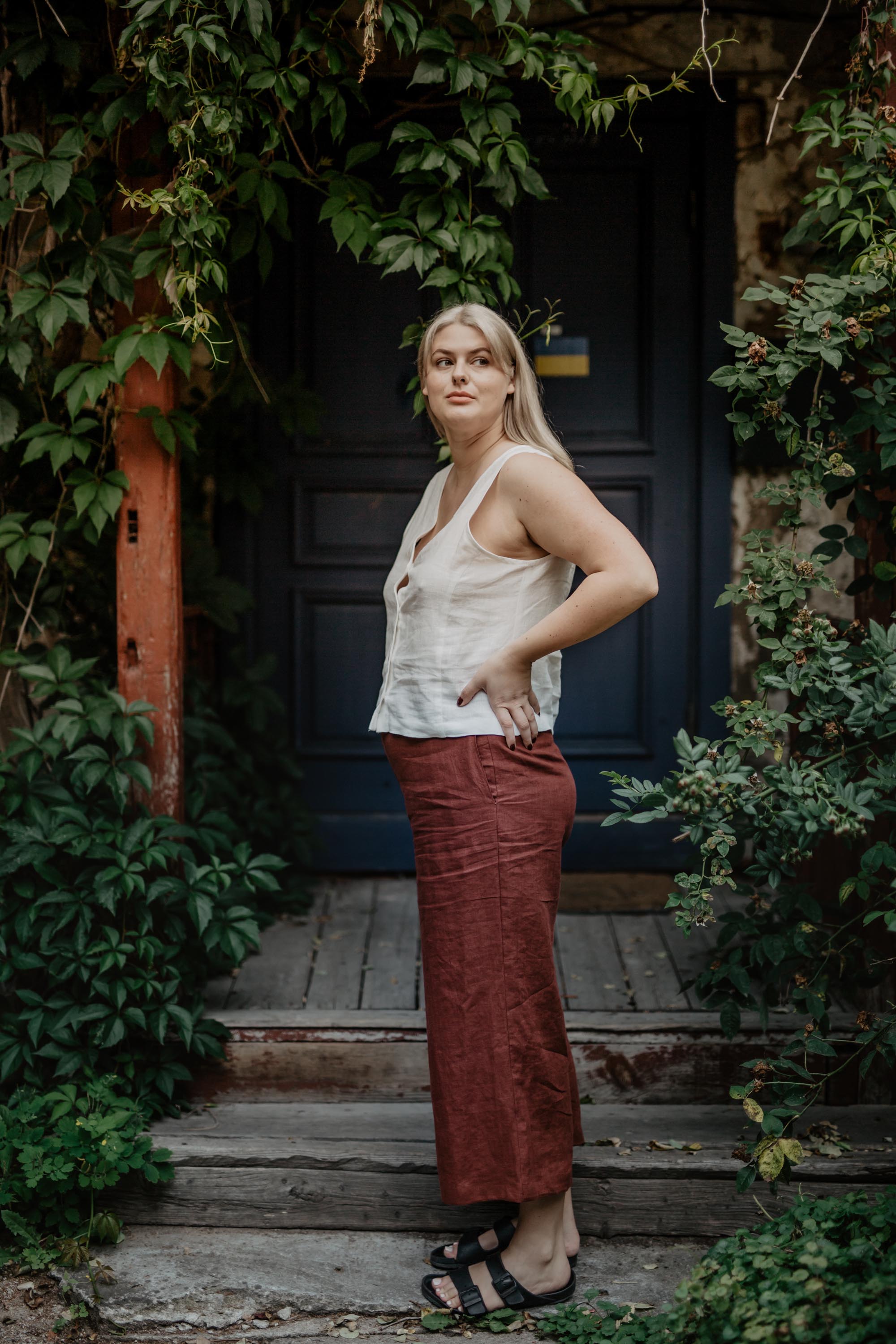 Woman In Looking To The Side In Botanical Setting Wearing A White Linen Button Top and Terracotta Pants By Amourlinen