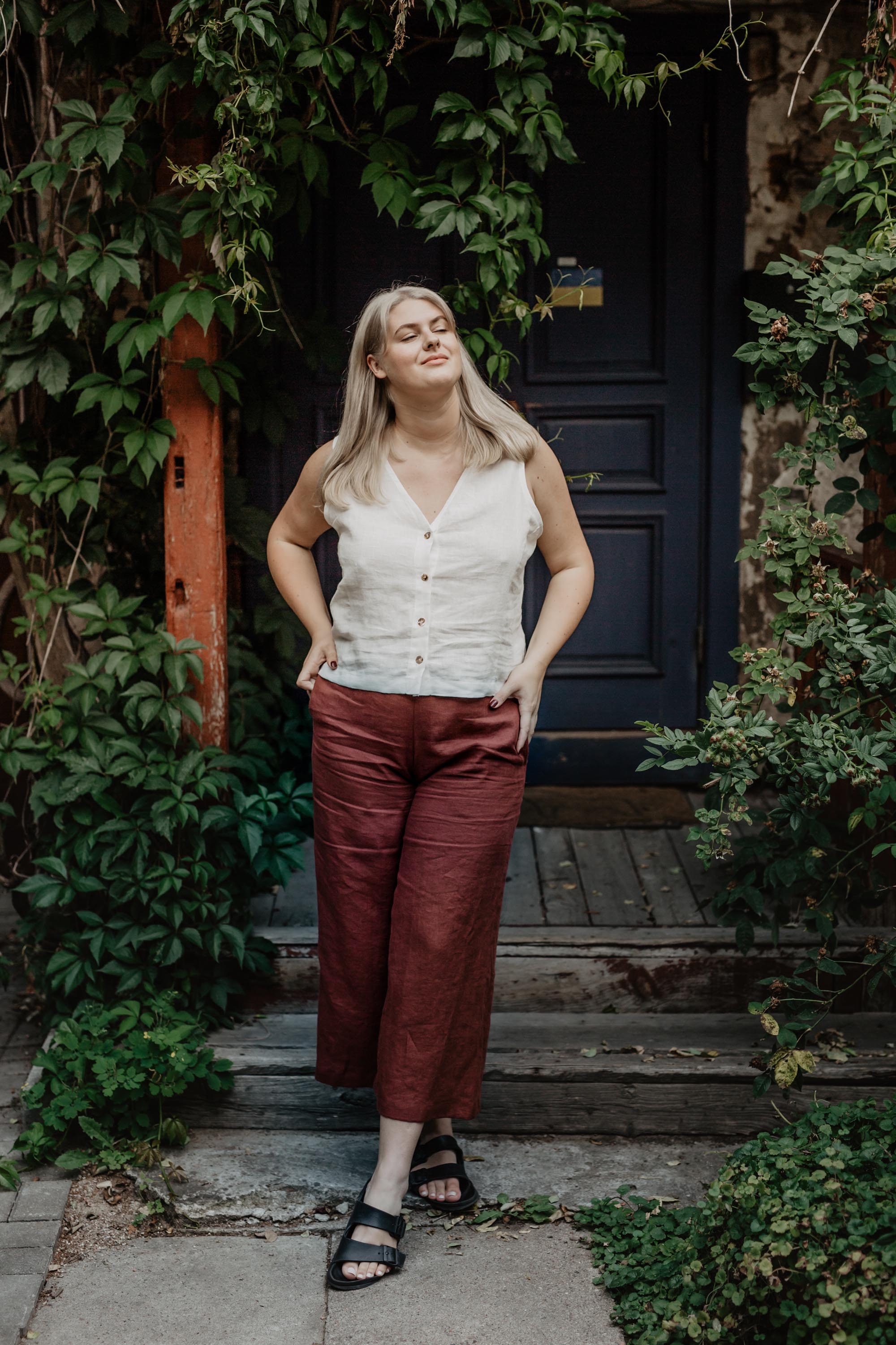 Woman In Botanical Setting Wearing A White Linen BUtton Top and Terracotta Linen Pants By Amourlinen