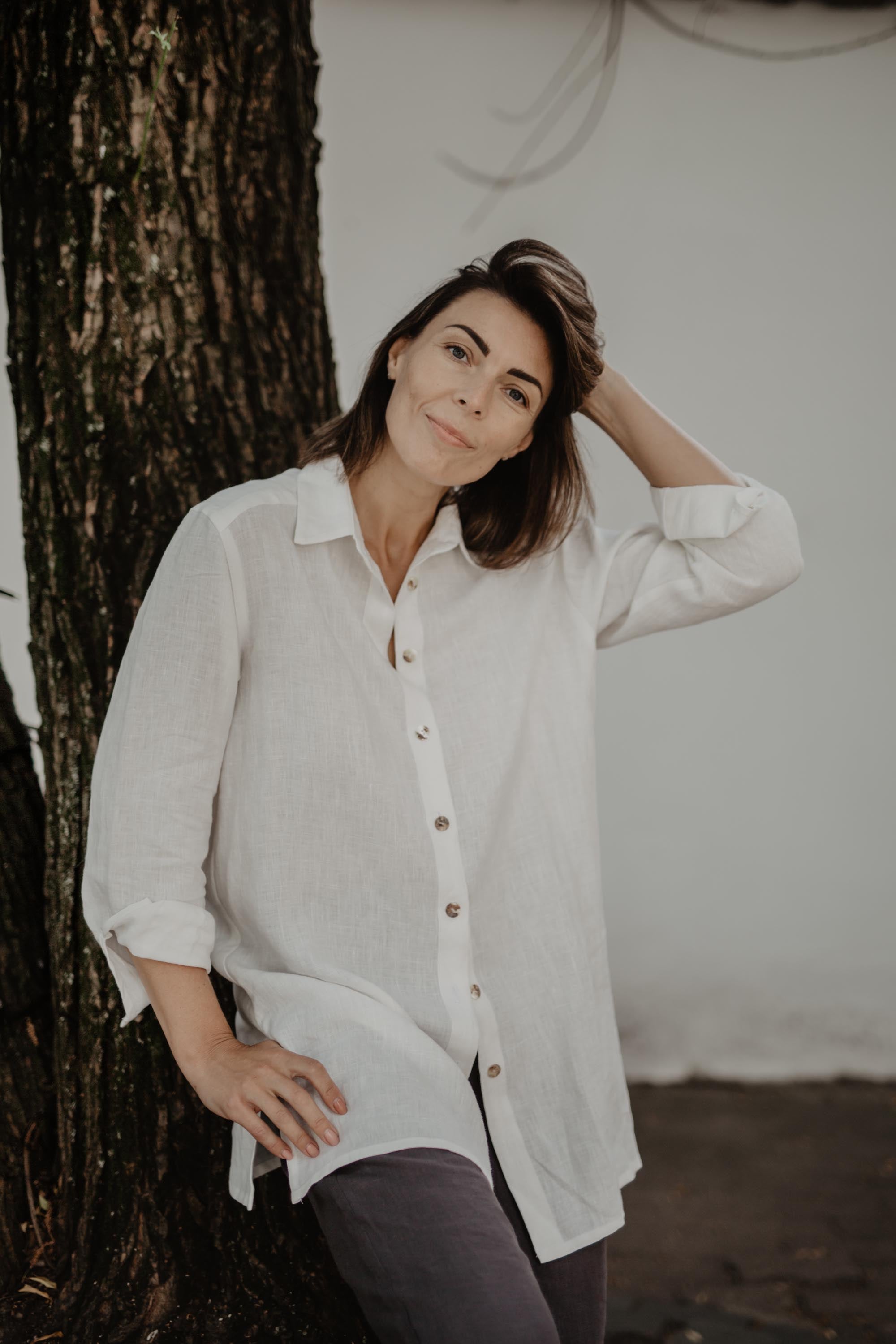 Woman Wearing A White Oversized LInen Shirt and Grey Pants Standing Next to A Tree