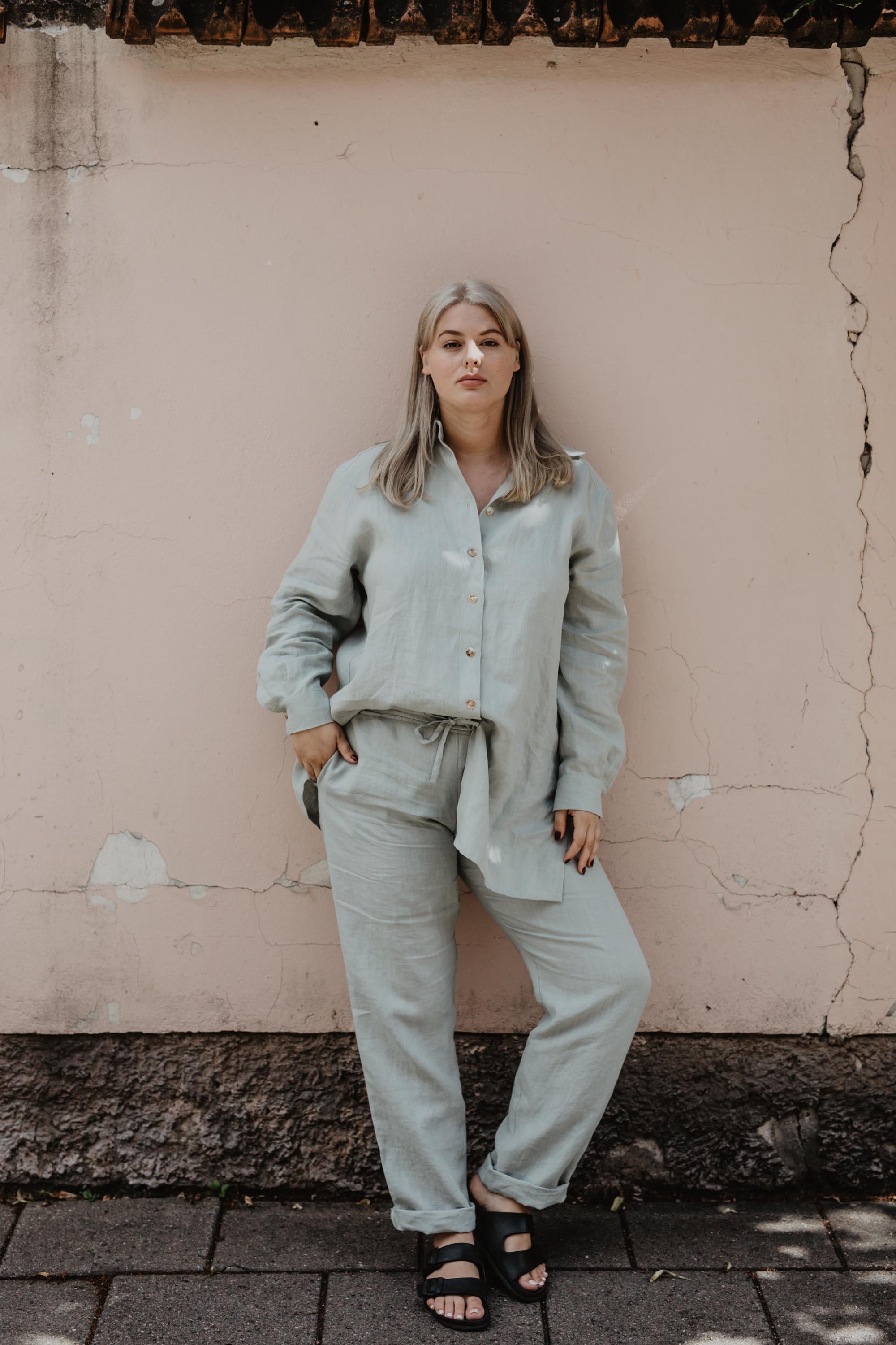 Woman Facing Forward Leaning Agains A Wall Wearing Sage Green Linen Suit Set By AmourLInen
